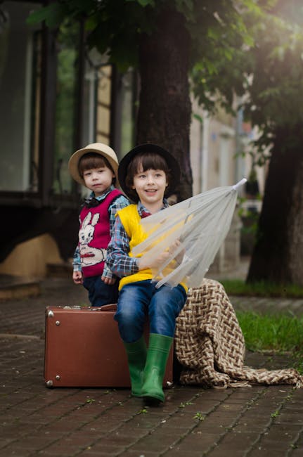 Two young boys sitting on a suitcase outdoors with an umbrella, exuding charm and adventure.