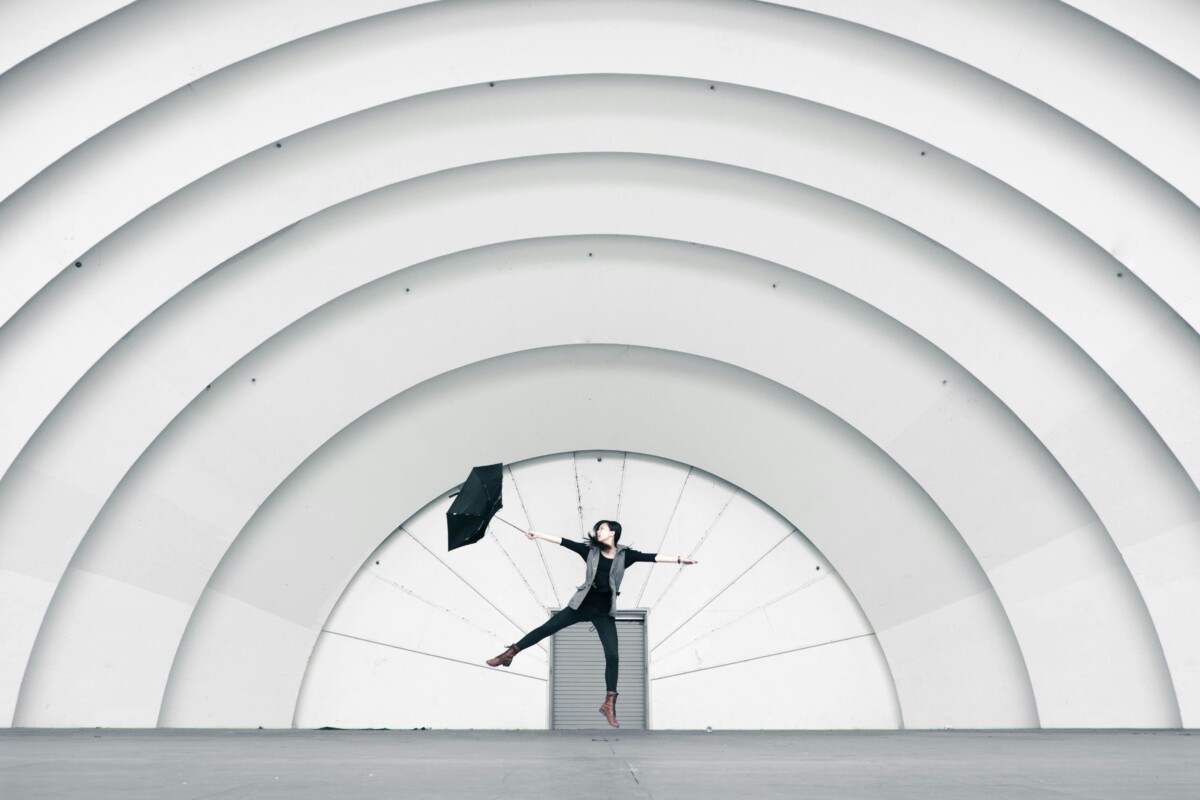 Person jumping joyfully under large architectural arches with an umbrella.